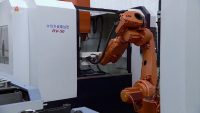 Image shows an orange industrial robot arm holding a piece of metal to a larger, white North Korean industrial machine in a factory